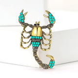 Scorpion brooch with gold-tone finish, teal and clear decorative stones, upright pose on white block.