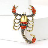 Scorpion brooch with gold-tone metal, orange-red and clear faceted stones, upright display on white block.