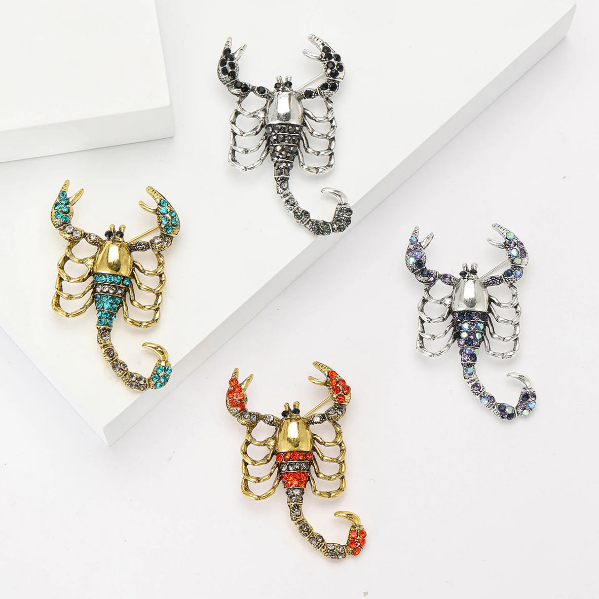 Set of four scorpion brooches with silver- and gold-tone finishes in black, teal, red, and iridescent blue stones, flat lay on white blocks.