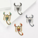 Set of four scorpion brooches with silver- and gold-tone finishes in black, teal, red, and iridescent blue stones, flat lay on white blocks.