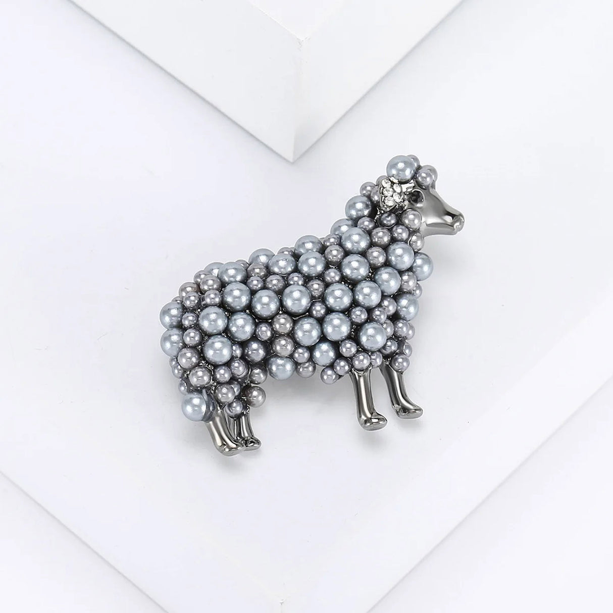 Sheep brooch with grey-tone metal and layered beads, angled top view on white platform.