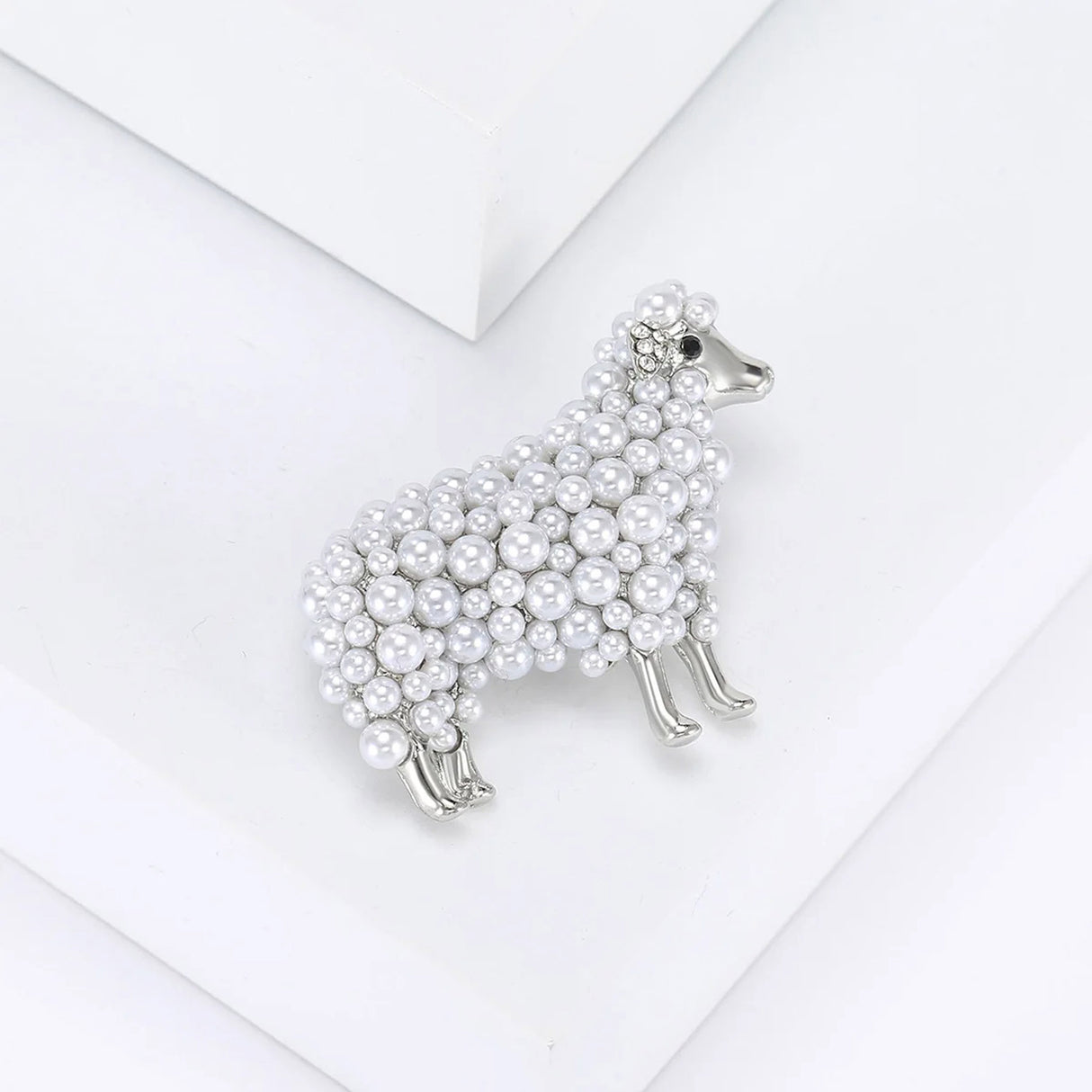 Sheep brooch with silver-tone metal, clustered white round beads, and small clear crystal ear accents, angled top view on white surface in studio.