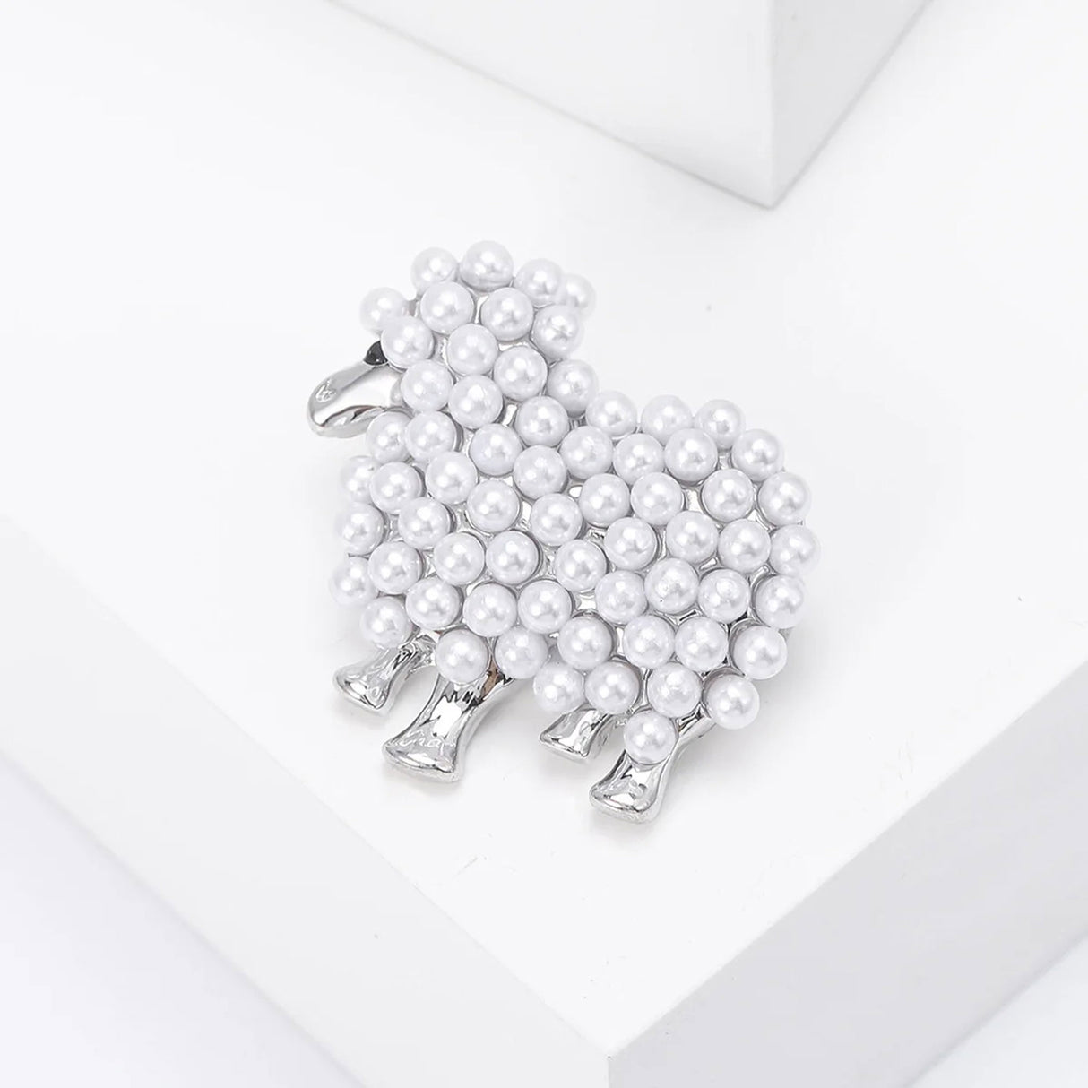 Sheep brooch in silver-tone metal with white round beads and black eye detail, angled top view on white studio surface.