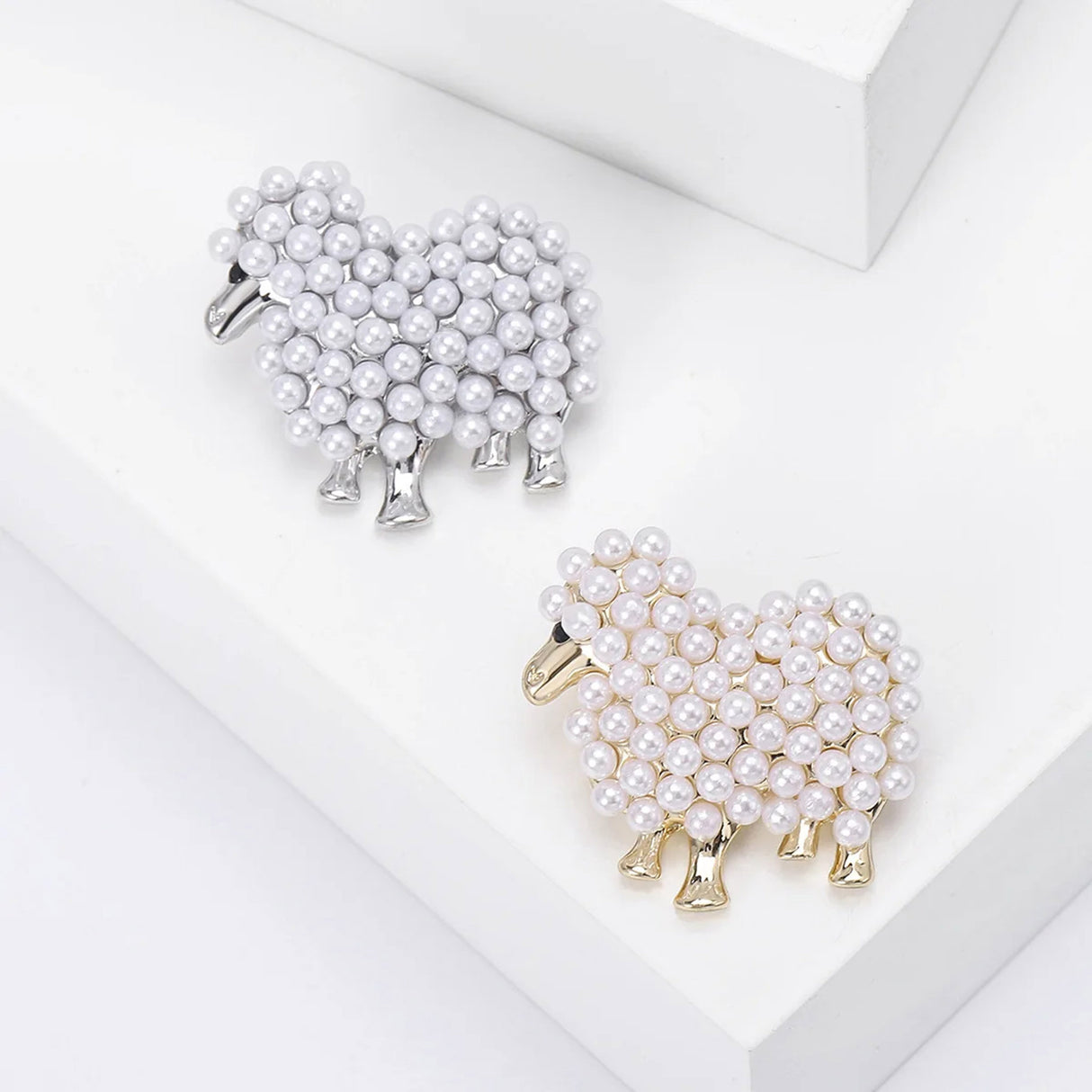 Two sheep brooches in silver-tone and gold-tone finishes with white round beads, top-down view on white background.