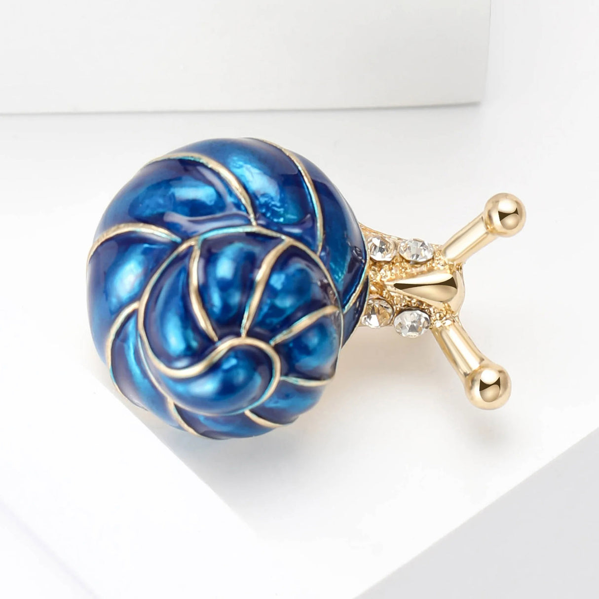 Snail brooch with blue-toned spiral shell, gold-tone body, and clear stones, angled flat lay on white.