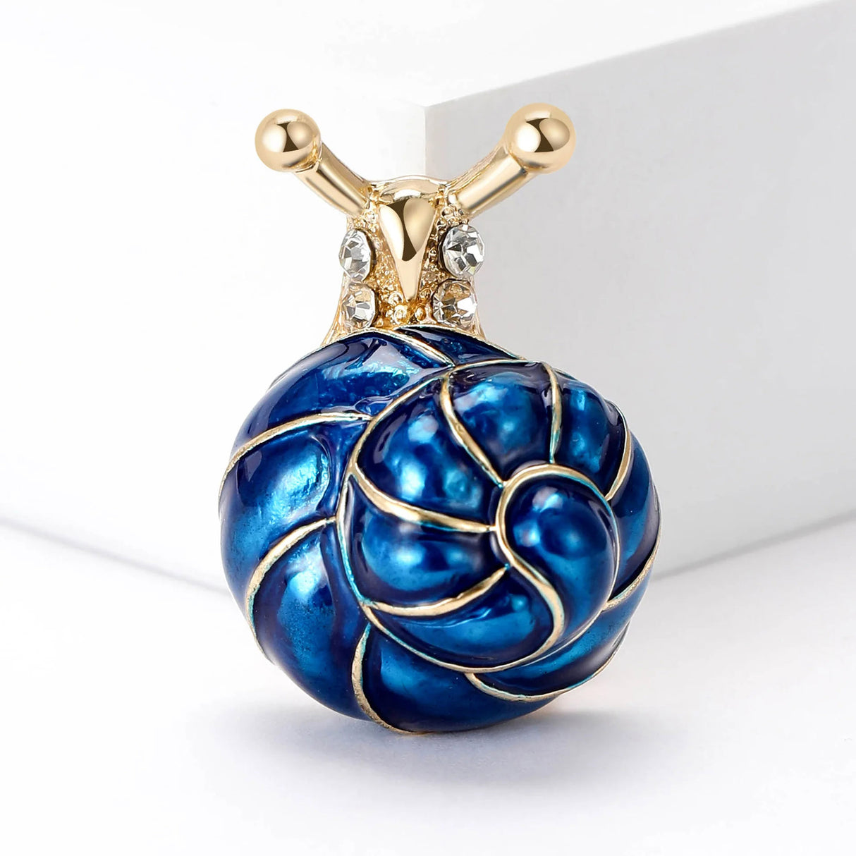 Snail brooch featuring blue-toned spiral shell, gold-tone body, and clear stones, upright view on white studio backdrop.