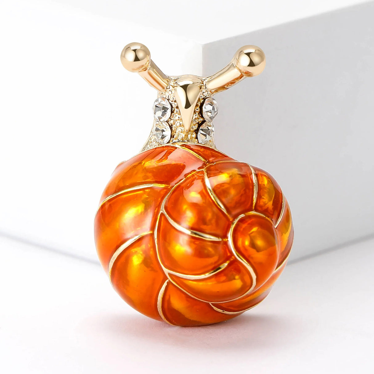 Snail brooch with orange-toned swirl shell, gold-tone metal and clear stones, upright studio view on white.
