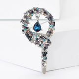 Snake brooch with silver-tone base, multicolor blue-purple-beige iridescent stones, and a hanging blue teardrop stone, upright on white block.