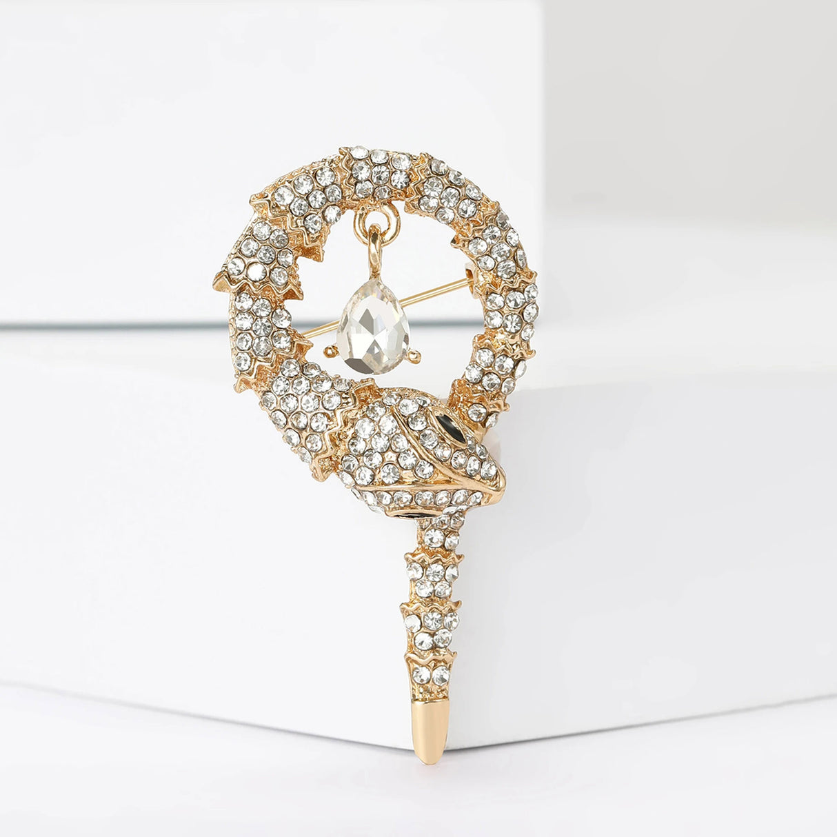 Snake brooch with gold-tone base, clear faceted stones, and a hanging clear teardrop stone, upright on white block.
