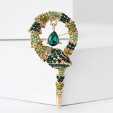 Snake brooch with gold-tone base, green and dark green faceted stones, and a hanging green teardrop stone, upright on white block.