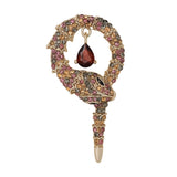 Snake brooch with gold-tone metal base, faceted stones in red, amber, and gray tones, and dark red teardrop pendant, cutout on white.