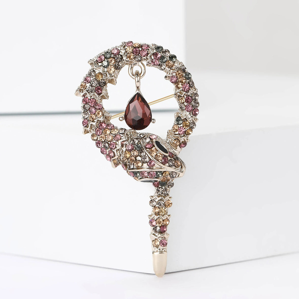 Snake brooch with gold-tone base, red and brown faceted stones, and a hanging red teardrop stone, upright on white block.