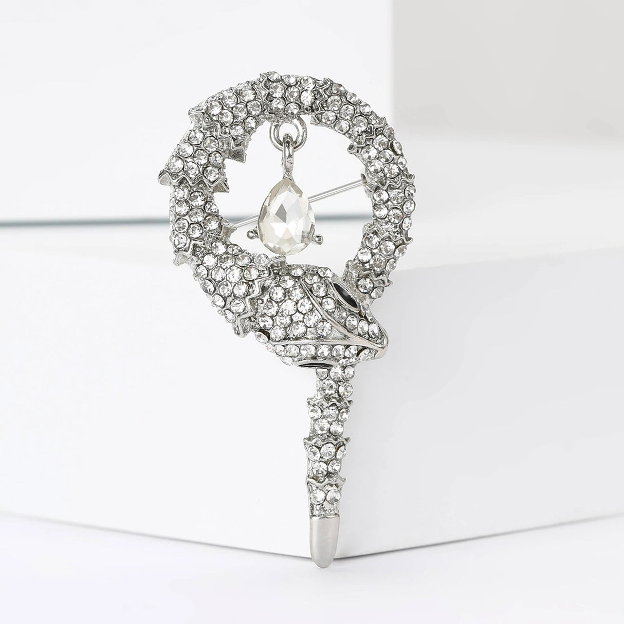 Snake brooch with silver-tone base, clear faceted stones, and a hanging clear teardrop stone, upright on white block.