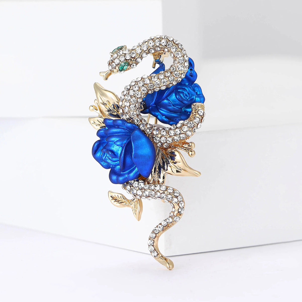 Snake brooch with clear round stones, metallic gold-tone leaves, and vivid blue rose motifs, upright pose on white.