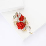 Snake brooch with clear faceted stones, green eyes, and bold red rose accents, top-down view on white surface.