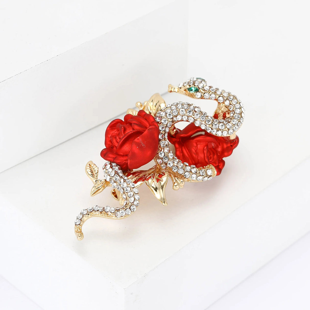 Snake brooch featuring clear stones, gold-tone leaves, and red sculpted roses, angled studio view on white block.