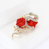 Snake brooch featuring clear stones, gold-tone leaves, and red sculpted roses, angled studio view on white block.