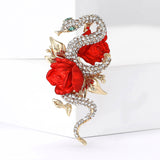 Snake brooch adorned with clear pave-set stones, gold-tone leaves, and two red rose motifs. Upright pose on white studio background.