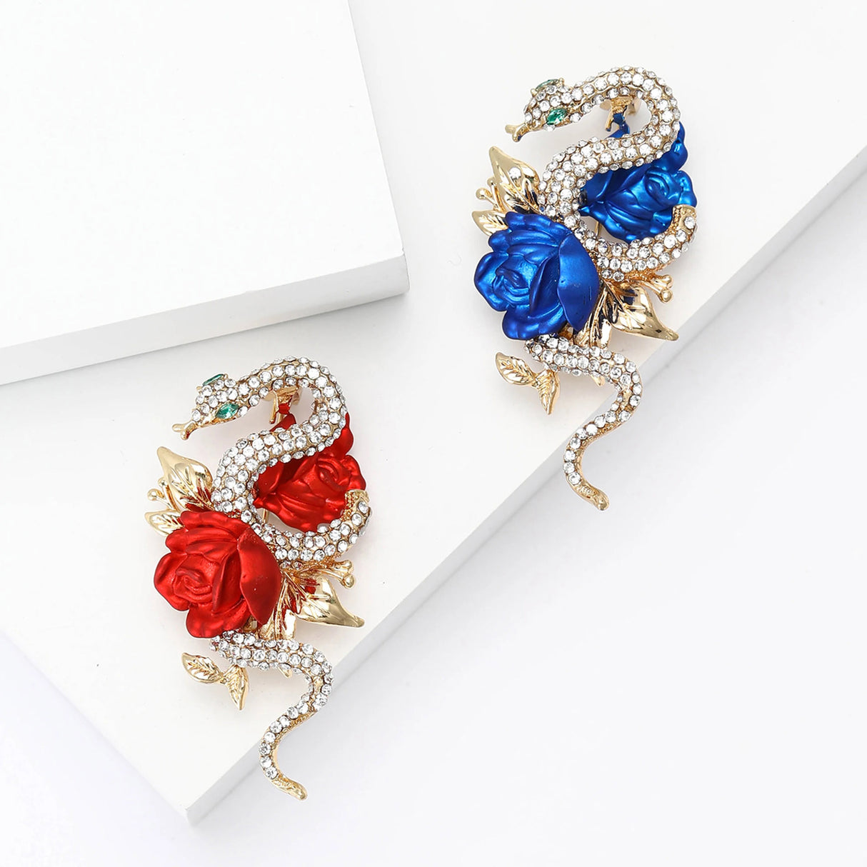 Pair of snake brooches in gold-tone metal, with red and blue roses and clear pave-set stones. Top-down view on white studio backdrop.