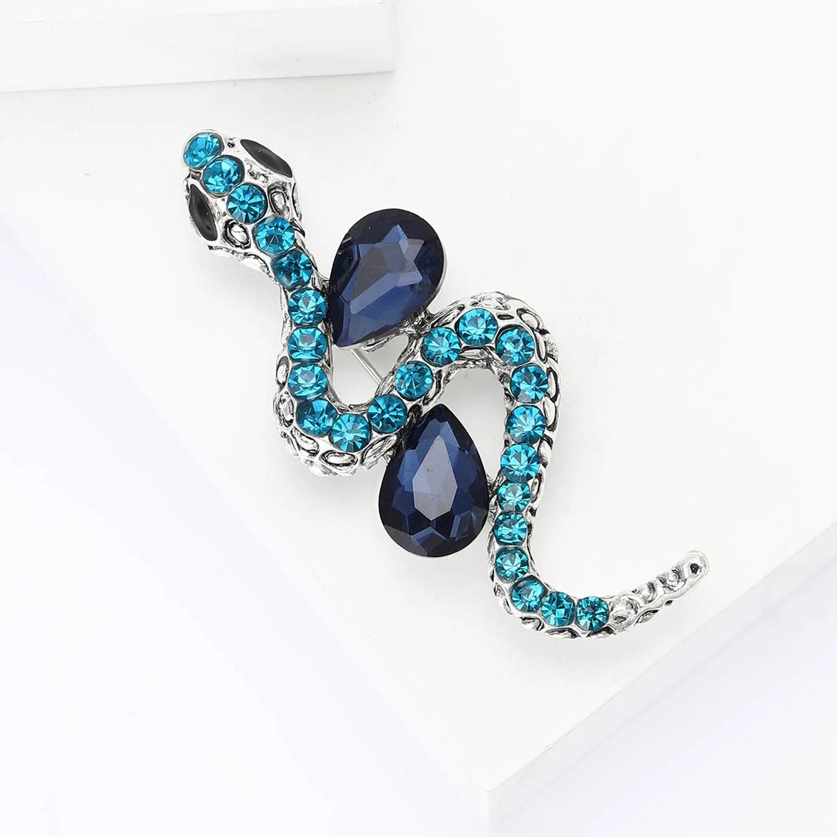 Snake brooch with silver-tone finish, deep blue faceted stones, and turquoise round crystals, top-down view on white.