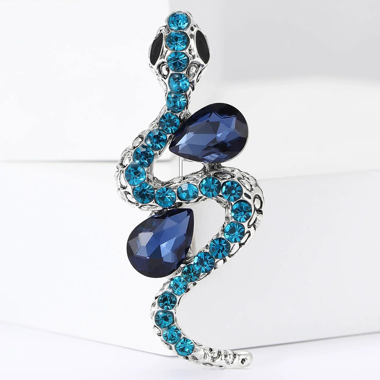 Snake brooch with silver-tone body, navy blue teardrop stones, and aqua blue crystals, upright pose on white block.