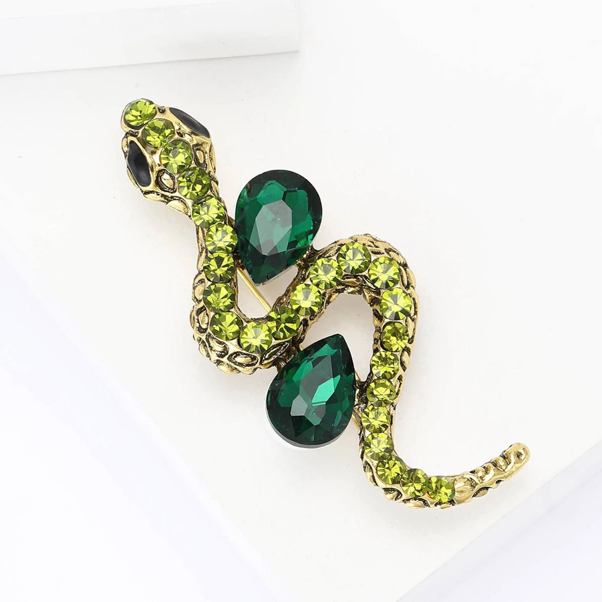 Snake brooch in gold-tone metal with dark green teardrop stones and yellow-green crystals, top-down view on white.