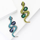 Pair of snake brooches in silver-tone and gold-tone metals with blue and green teardrop stones, flat lay on white blocks.