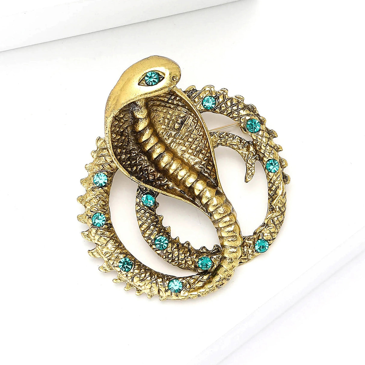 Gold-tone cobra brooch with green faceted accent stones and textured scales, angled flat lay on white studio surface.