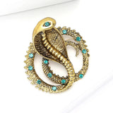 Gold-tone cobra brooch with green faceted accent stones and textured scales, angled flat lay on white studio surface.