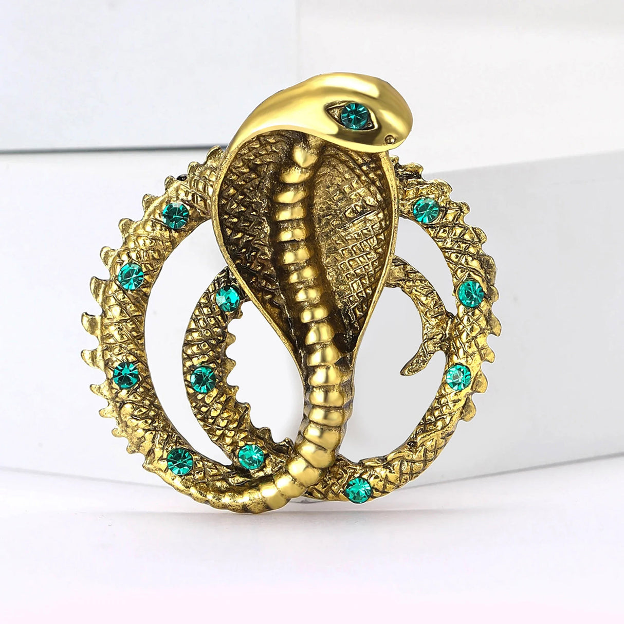Cobra snake brooch in gold-tone metal finish with green decorative stones and textured coiled body, upright on white surface.