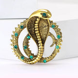 Cobra snake brooch in gold-tone metal finish with green decorative stones and textured coiled body, upright on white surface.