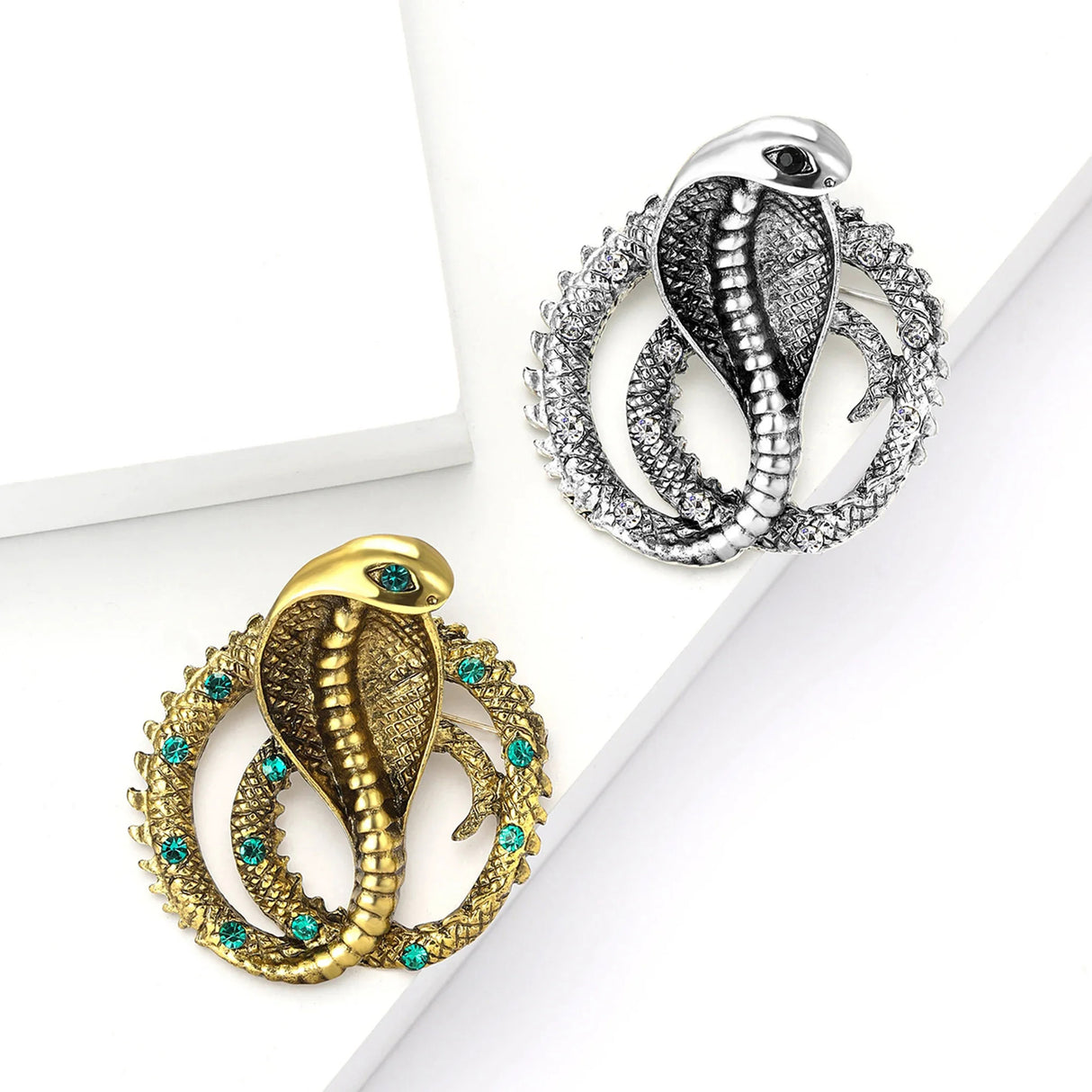 Set of two cobra-shaped snake brooches in gold-tone with green stones and silver-tone with clear stones, flat lay on white blocks.