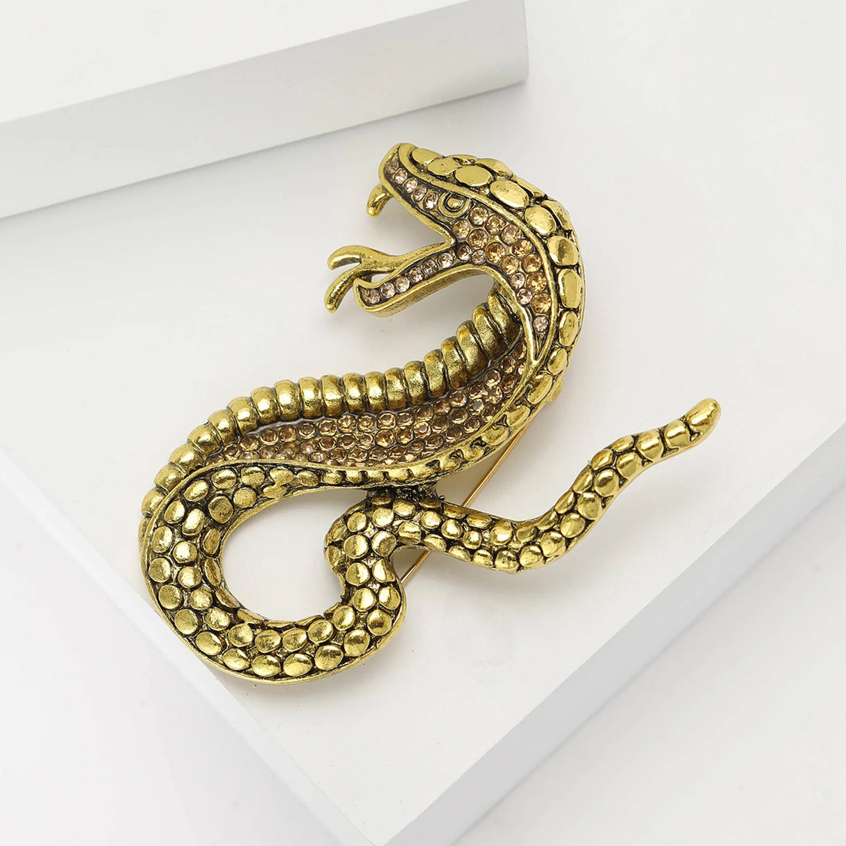 Gold-tone snake brooch with textured scales and small light brown stones, flat lay on white surface.
