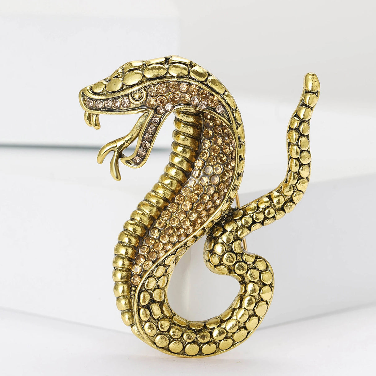 Snake brooch in gold-tone metal with textured scale pattern and light brown faceted stones, standing on white platform.