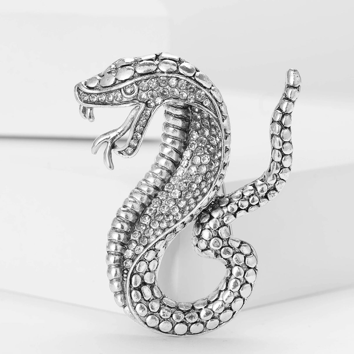 Silver-tone snake brooch with clear faceted stones and textured scale details, upright pose on white block.