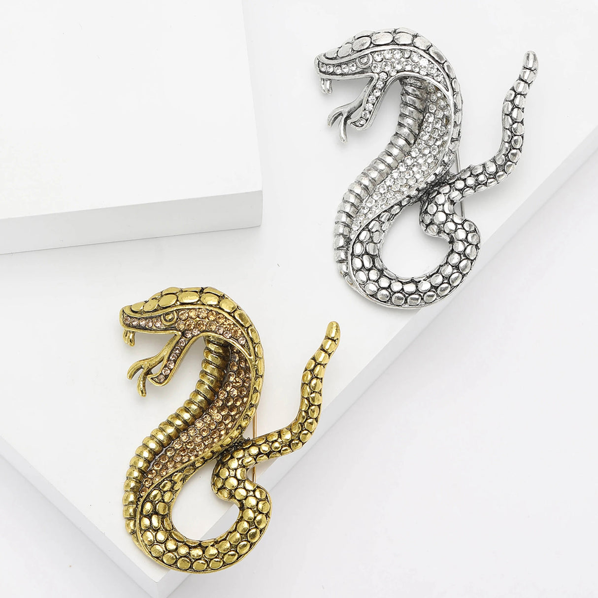 Pair of snake brooches in gold-tone and silver-tone metal finishes with clear and brown round accent stones. Flat lay on white blocks in studio.