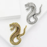 Pair of snake brooches in gold-tone and silver-tone metal finishes with clear and brown round accent stones. Flat lay on white blocks in studio.