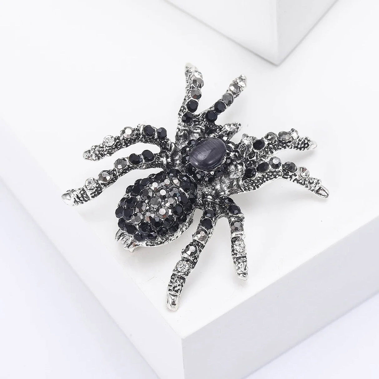 Tarantula brooch with silver-tone finish, black and clear stones, and a dark oval cabochon, angled view on white block.