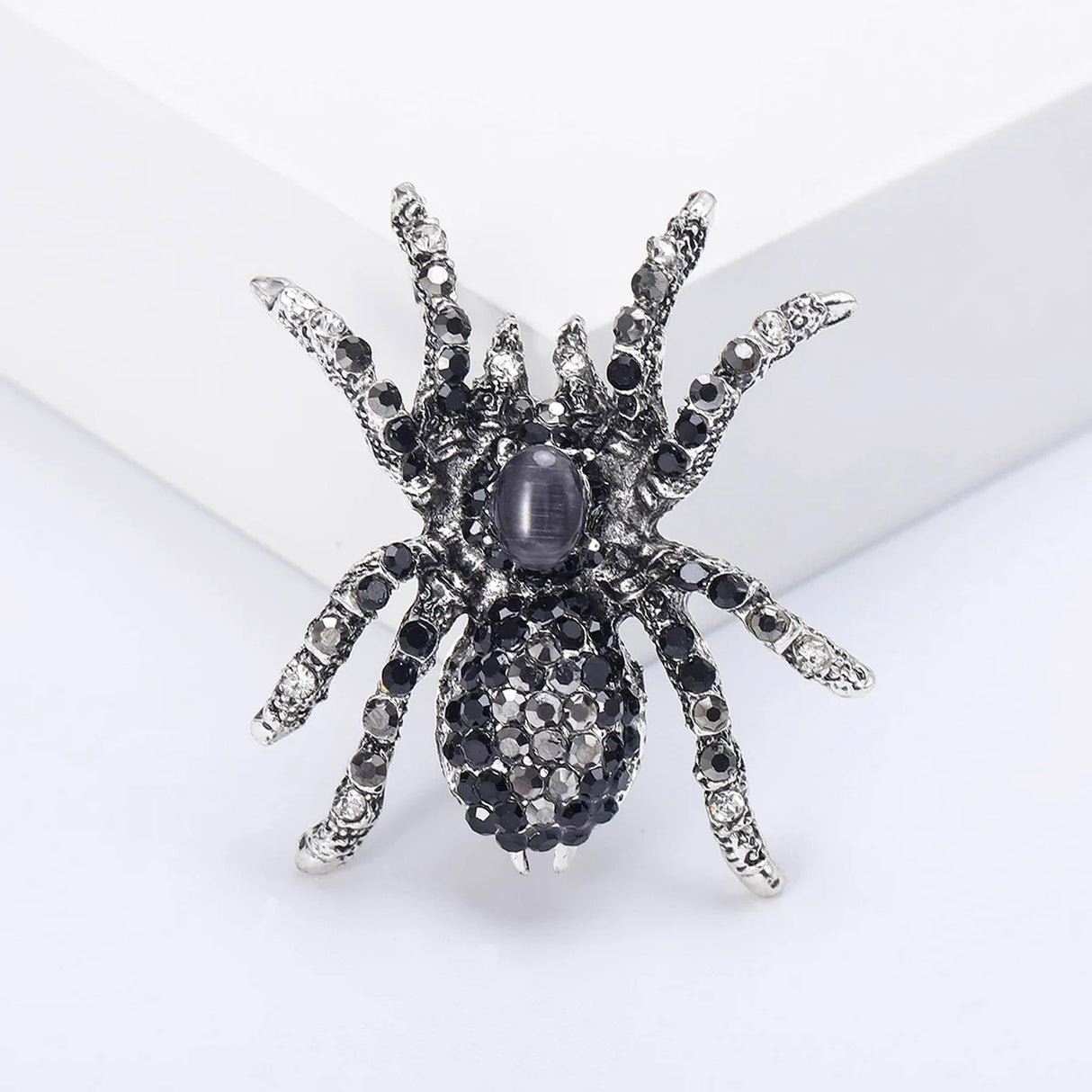 Spider brooch with black faceted stones, clear accents, and a dark oval cabochon center, upright view on white block.