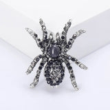 Spider brooch with black faceted stones, clear accents, and a dark oval cabochon center, upright view on white block.