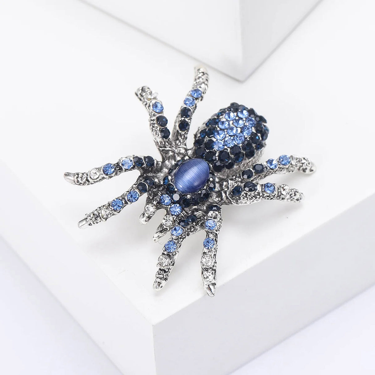 Tarantula brooch with silver-tone base, blue faceted stones, and a blue oval cabochon center, angled view on white block.