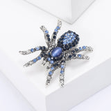 Tarantula brooch with silver-tone base, blue faceted stones, and a blue oval cabochon center, angled view on white block.