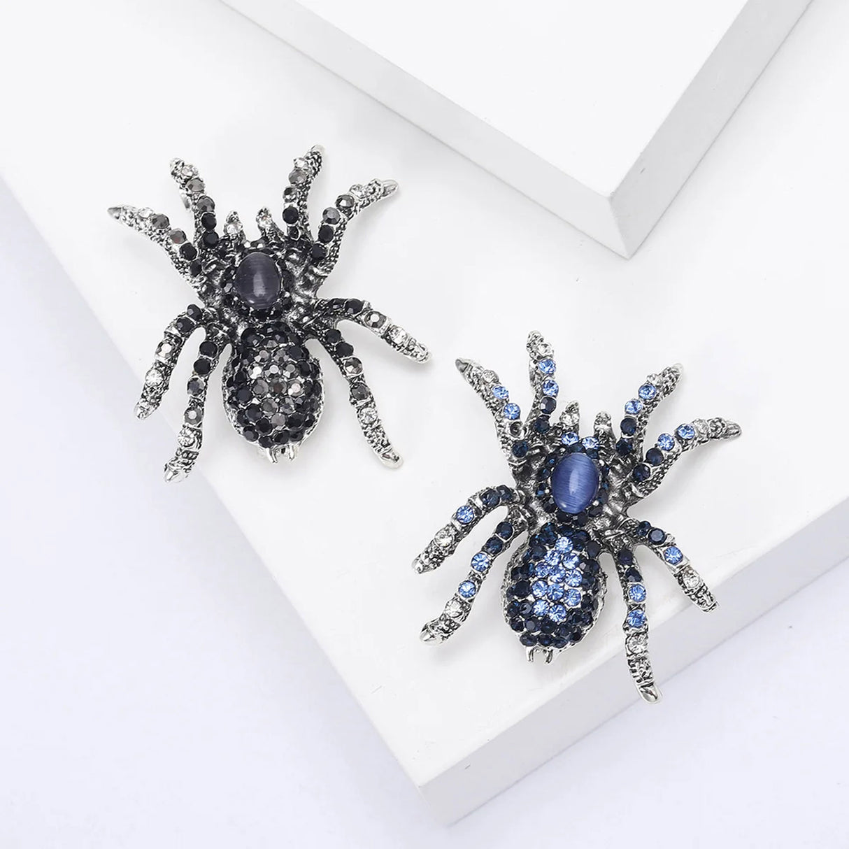 Pair of spider brooches with silver-tone bases, one in black and clear stones, the other in blue tones, flat lay on white platform.