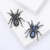 Pair of spider brooches with silver-tone bases, one in black and clear stones, the other in blue tones, flat lay on white platform.