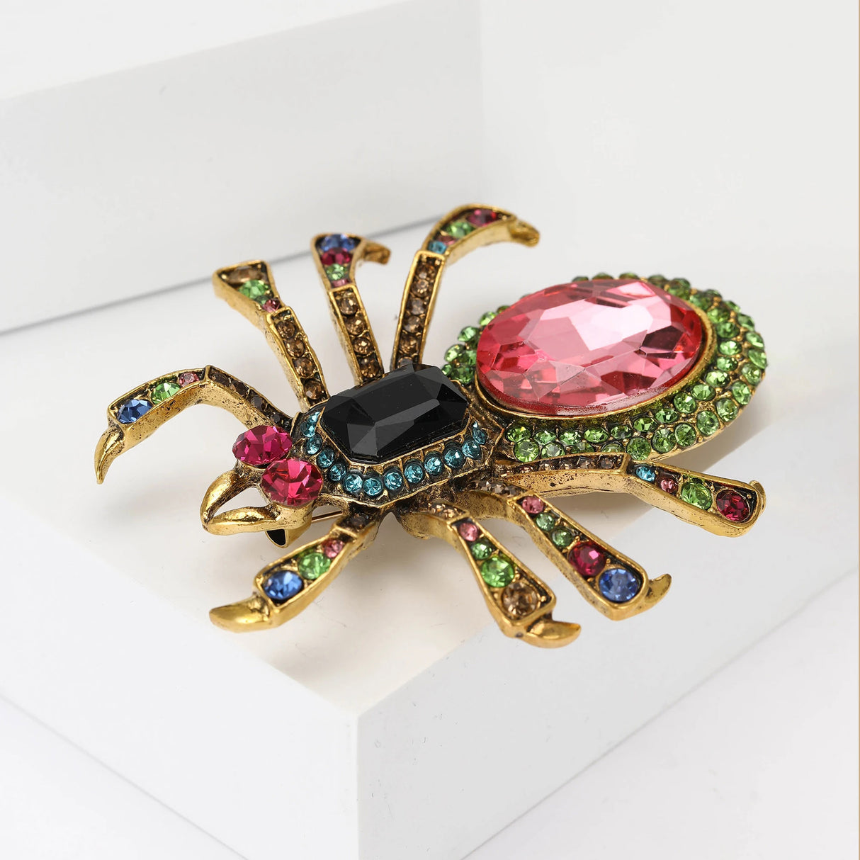 Spider brooch with gold-tone metal base, large pink faceted stone, black center gem, and multicolor accents, angled view on white platform.