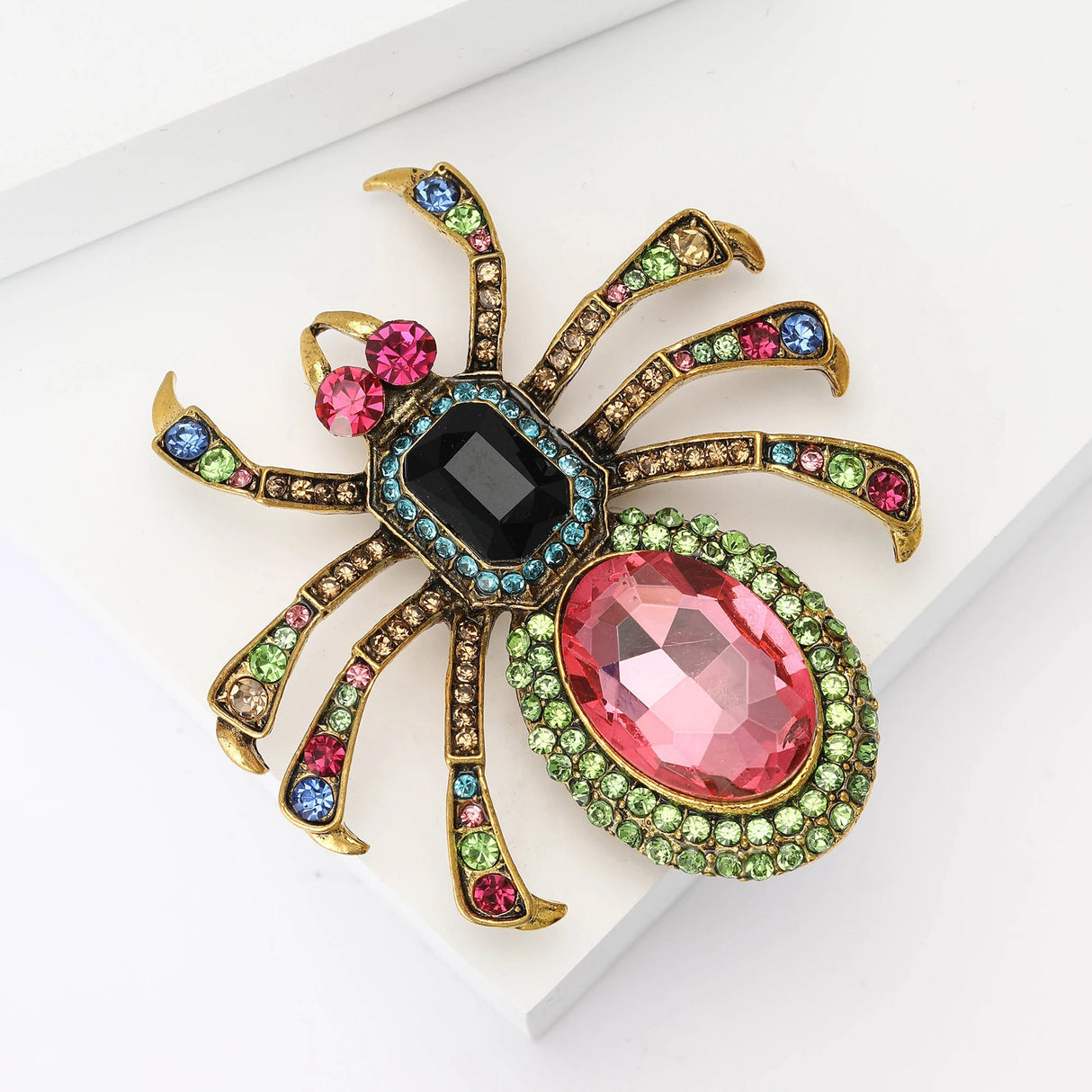 Spider brooch with pink oval stone bottom, black rectangular stone body, and multicolor faceted details, flat lay on white surface.
