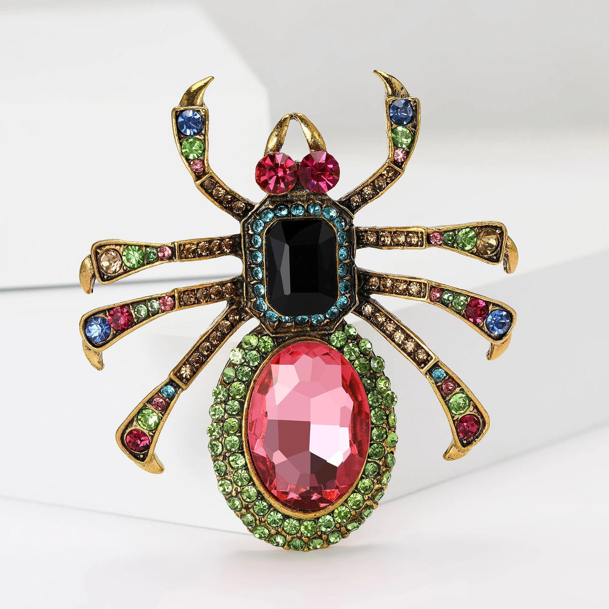 Spider brooch with gold-tone metal legs, pink oval stone bottom, black square stone body, and multicolor embellishments. Upright pose on white studio backdrop.
