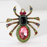 Spider brooch with gold-tone metal legs, pink oval stone bottom, black square stone body, and multicolor embellishments. Upright pose on white studio backdrop.