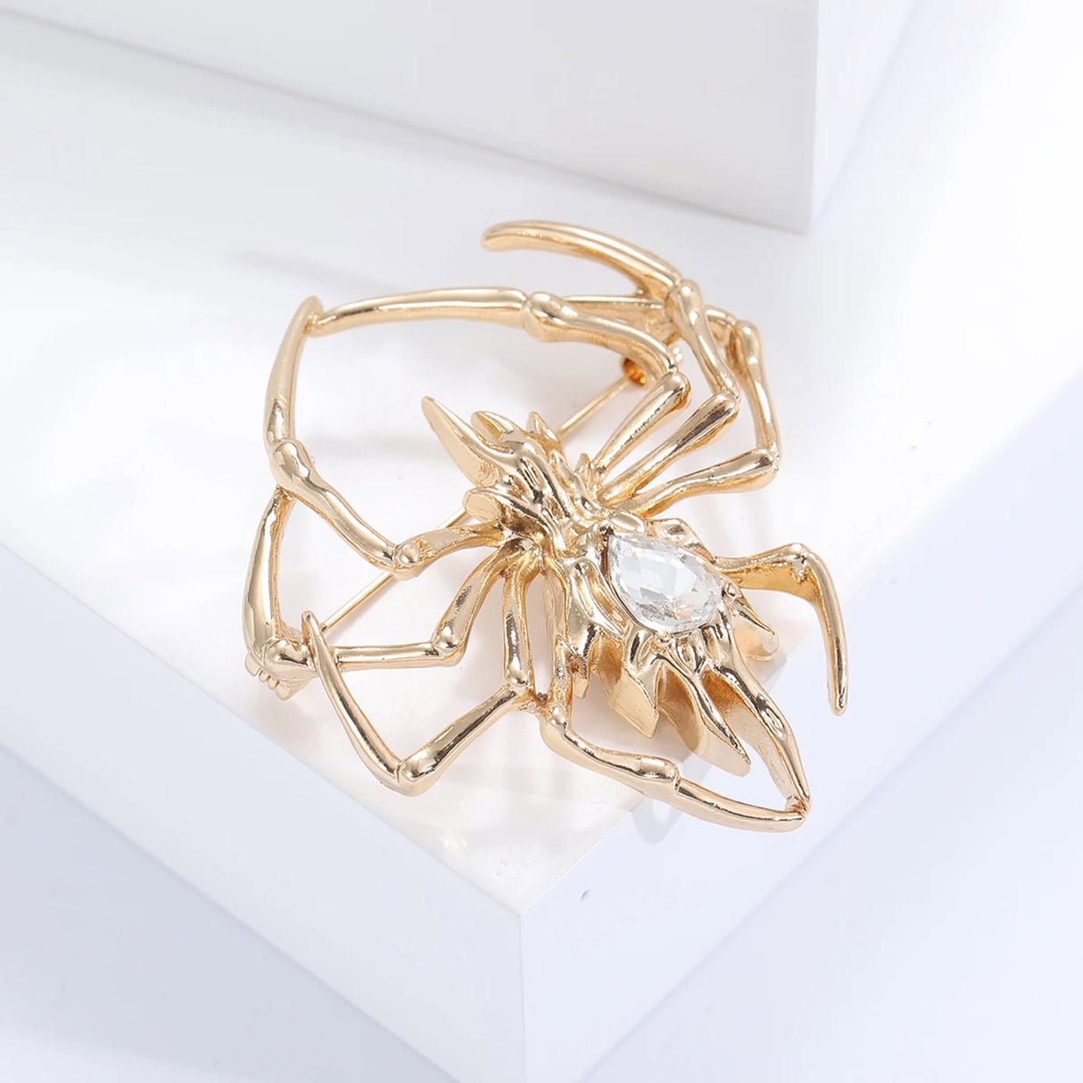 Gold-tone spider brooch with clear teardrop stone and textured legs, side view from top on white block.