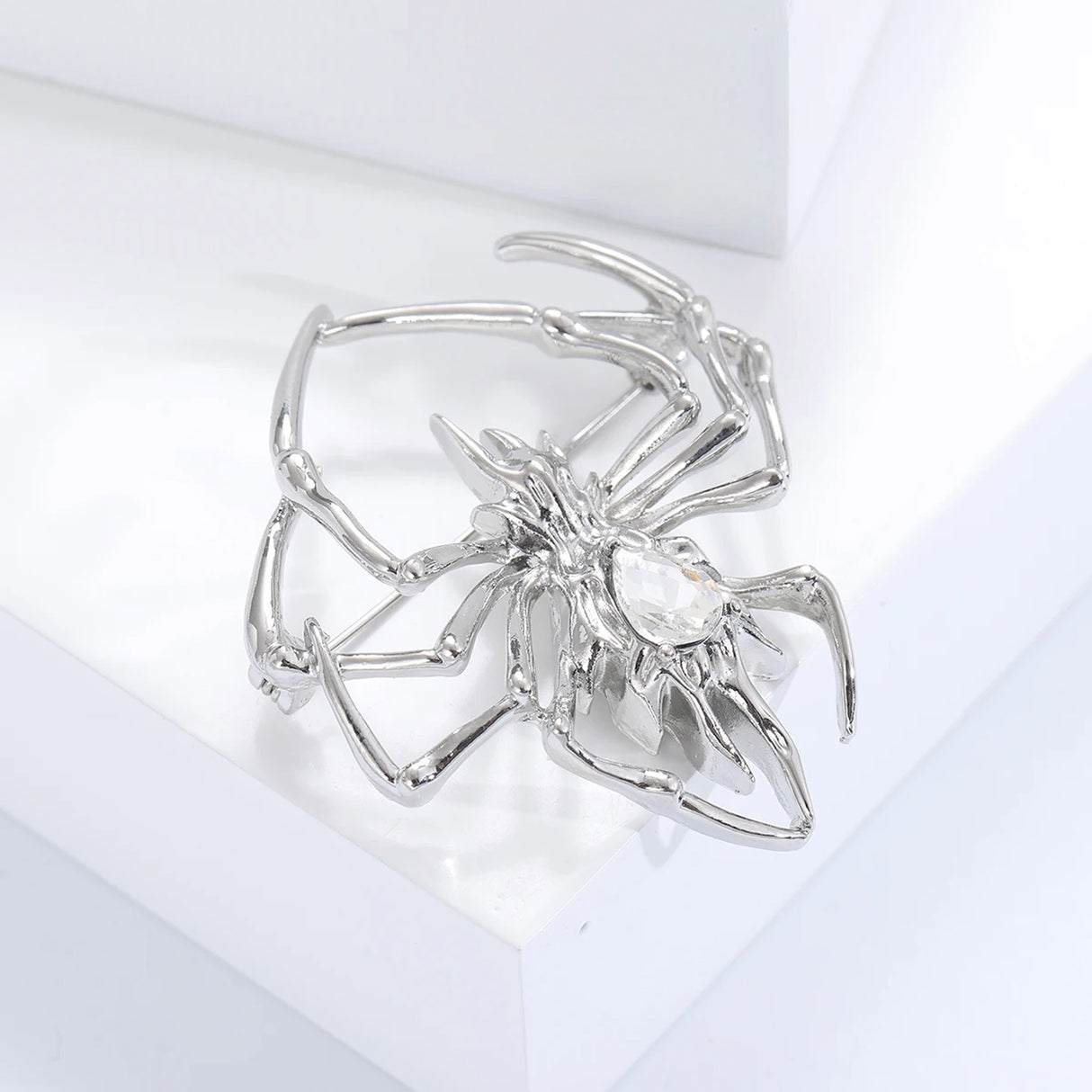 Spider brooch in polished silver-tone metal with large clear faceted teardrop center stone, side view on white block.